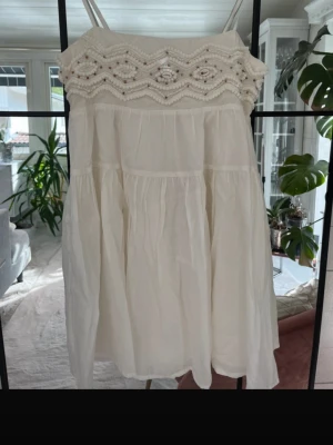 Vit kort klänning med pärldetaljer - Söt vit kortklänning med smala axelband och vacker pärlbroderad överdel. Klänningen har en luftig passform och är tillverkad i ett lätt tyg, perfekt för varma dagar. Den har en bohemisk känsla och volangliknande fall.