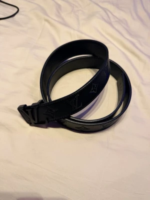 Svart bälte i skinn - Snyggt svart bälte i skinn med klassiskt monogrammönster präglat längs hela bältet. Bältet har en stilren svart spänne som matchar resten av designen. Perfekt accessoar för att lyfta din outfit.