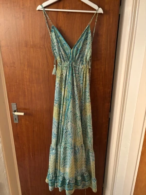 Långklänning med blå/grönt mönster - Säljer en somrig långklänning med tunna axelband och v-ringning. Klänningen har ett blått och grönt mönster som påminner om blad, samt volangkant nertill. Materialet känns lätt och luftigt, perfekt för varma dagar. 