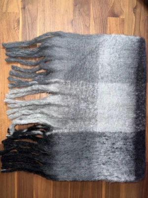 Rutig halsduk i svart och grått - Mjuk och fluffig halsduk med rutigt mönster i svart och grått. Halsduken har långa fransar i båda ändar och är perfekt för att hålla värmen under kalla dagar. Materialet känns ulligt och ger en mysig känsla. Halsduken liknar ”Acne Studios halsdukarna och är oanvänd (ny).