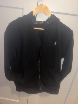 Svart hoodie från Polo Ralph Lauren - Svart hoodie från Polo Ralph Lauren med dragkedja framtill, huva med snörning och klassisk logga broderad på bröstet. Mjuk och skön, perfekt för en avslappnad stil. Fickor framtill och ribbade muddar vid ärmslut och nederkant.