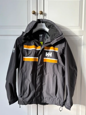 Svart regnjacka Helly Hansen M - Svart regnjacka från Helly Hansen med huva och dragkedja. Jackan har vita och gula ränder på bröstet och ryggen samt HH-logga fram och bak. Tillverkad i vattenavvisande material och har justerbar nederkant och muddar. Perfekt för regniga dagar.