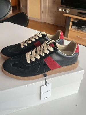 Maison Margiela Gats - Snygga svarta sneakers med röd sidodetalj och beige innersula. Skorna har beigea skosnören och en klassisk gummisula i brunt. Materialet ser ut att vara mocka och läder, vilket ger en stilren och trendig look.