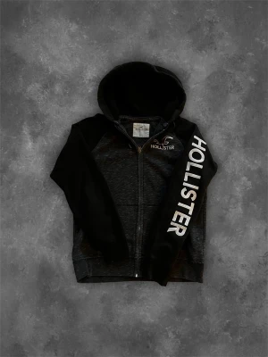 Svart zip hoodie från Hollister - Snygg svart hoodie från Hollister med dragkedja och stor huva. Stora vita HOLLISTER-tryck på ena ärmen och mindre logga på bröstet. Tillverkad i mjukt material som känns skönt mot huden. Perfekt för en avslappnad streetwear-look. 