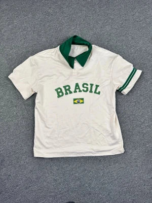 Vit Brasil t-shirt med grön krage - Vit t-shirt med grön krage och gröna detaljer på ena ärmen. På bröstet står det 'BRASIL' och en liten brasiliansk flagga är tryckt under texten. Perfekt för dig som gillar sportig stil och vill visa ditt stöd för Brasilien.