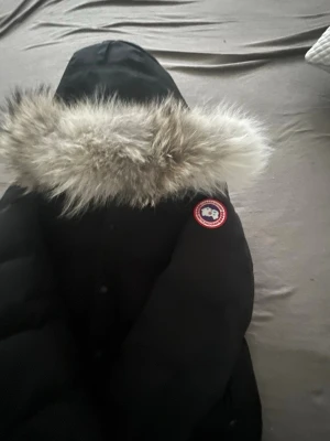 Svart parkas från Canada Goose - Säljer en svart parkas från Canada Goose med stor huva och fluffig pälsdetalj. Jackan har klassisk logotyp på ärmen och är vadderad för extra värme. Perfekt för kalla dagar och riktigt snygg till vintern.
