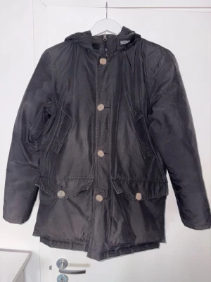  Woolrich Down Parka Jacket - Säljer en svart parkas från Woolrich med huva och stora knappar framtill. Jackan har två snedställda bröstfickor och två stora fickor med lock nedtill. Insidan är beige och jackan har en klassisk, rak passform. Perfekt för kalla dagar. Såklart Äkta och Nyligen kemtvättad för någon månad sen