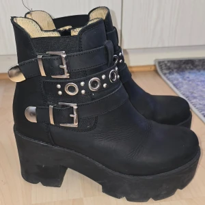 Svarta boots Johnny Bulls - Säljer ett par svarta boots med grov platåsula och högt skaft. Skorna har två breda spännen i silver, dekorativa nitar och ringar runt ankeln samt detaljer i metall. 