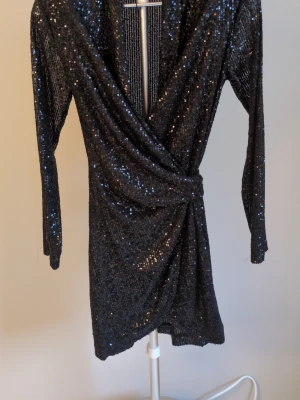 Svart glittrig omlottklänning - Snygg svart kort omlottklänning med långa ärmar och paljettdetaljer som ger ett glittrigt intryck. Klänningen har ett knyt i midjan och djup v-ringning framtill. Perfekt för dig som vill sticka ut med en festlig look. Paljetter har lossnat på ena axeln, se bild ☺️