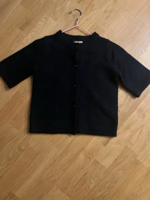 Svart kofta med kort ärm - En svart kofta med rund halsringning och knäppning framtill. Koftan har korta ärmar och rak passform, perfekt att slänga över en t-shirt eller linne. Enkel och stilren design som funkar till många olika looks.