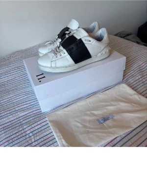 Vit-svarta sneakers från 11 by Boris Bidjan - Snygga vita sneakers med bred svart rem över foten från 11 by Boris Bidjan Saberi. Klassisk låg modell med vita snören och gummisula. Skorna har en modern och stilren design som sticker ut med den svarta detaljen.