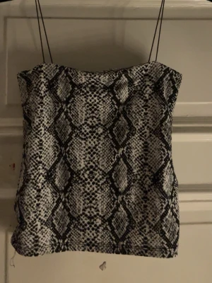 Tubtopp med ormskinnsmönster - Säljer en snygg tubtopp med svartvitt ormskinnsmönster och tunna axelband. Toppen har en rak passform och är perfekt för dig som vill sticka ut. Materialet känns mjukt och följsamt mot huden.