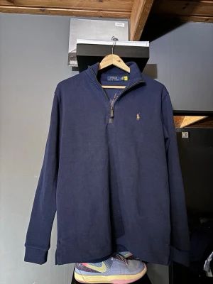 Mörkblå halvzip från Polo Ralph Lauren - Mörkblå långärmad tröja från Polo Ralph Lauren med halv dragkedja och klassisk broderad logga på bröstet. Tröjan har hög krage och ribbade muddar. Perfekt för en stilren och avslappnad look.