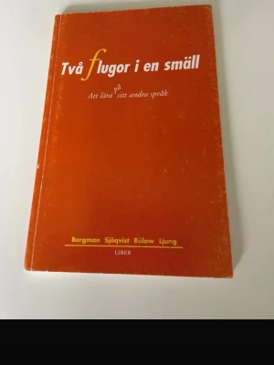 Två flugor i en smäll kursbok - Kurslitteratur med orange mjukt omslag, titeln 'Två flugor i en smäll'. Boken handlar om att lära sig sitt andra språk och innehåller teori, praktik och exempel för undervisning i svenska som andraspråk. Författare: Bergman, Sjöqvist, Bülow, Ljung.