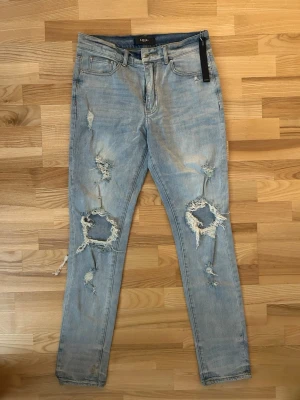 Ljusblå slitna jeans från Amiri  - Säljer ett par ljusblå jeans från Amiri med kraftiga slitningar och hål på båda benen. Modellen har fem fickor, normal midja och raka ben. Perfekta för en avslappnad streetstil. Materialet är klassisk denim i bomull.