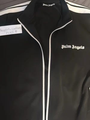 Palm Angels svart tracktröja - Svart kofta/ jacka från Palm Angels med vit logga broderad på bröstet. Jackan har dragkedja och ståkrage, samt vita ränder längs axlarna. Perfekt för en sportig och trendig streetwear-look. Strl M/L.  
