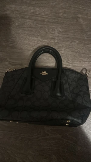 Svart handväska från Coach - Snygg svart handväska från Coach med klassiskt monogrammönster och guldfärgade detaljer. Väskan har två stadiga handtag, dragkedja upptill och är tillverkad i canvas med läderdetaljer. Perfekt för dig som gillar stilrena accessoarer.