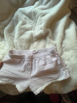 Ljusrosa pyjamasshorts med spetskant - Säljer ett par ljusrosa pyjamasshorts från Gina Shortsen har dekorativa knappar framtill, elastisk midja och söt spetskant längst ner, är i storlek 134/140 barn,tajt passform.