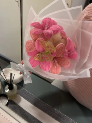Bukett med tygblommor och fjäril - Säljer en unik bukett med handgjorda tygblommor i rosa och aprikos nyanser, dekorerad med en dekorativ fjäril i gult och rosa. Buketten är inslagen i transparent papper och knuten med ett vitt band. Perfekt som accessoar eller dekoration.