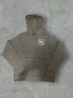 Grå hoodie från Hollister - Snygg grå hoodie från Hollister med vit logga på bröstet. Klassisk känguruficka framtill och huva. Mjuk och skön bomullsblandning, perfekt för en avslappnad stil.