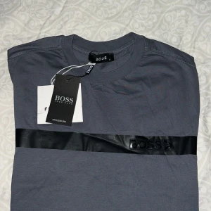 Grå BOSS t-shirt med svart detalj - Snygg grå t-shirt från BOSS med en svart glansig rand över bröstet och BOSS-loggan tryckt på bandet. Klassisk rund halsringning och korta ärmar. Perfekt för dig som gillar stilrena plagg med en twist.