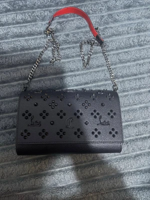 Svart kuvertväska från Louboutin - Snygg svart kuvertväska från Christian Louboutin i läder med dekorativa nitar och blommönster på framsidan. Väskan har en silverfärgad kedjerem och en röd detalj på handtaget, samt magnetlås. Perfekt för dig som vill sticka ut med en exklusiv accessoar.