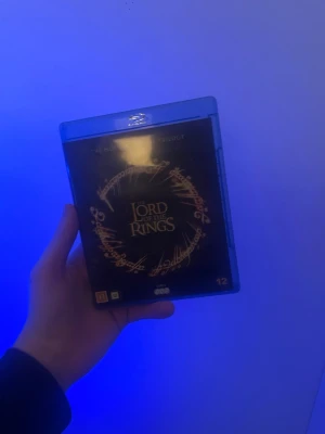 Lord of the Rings Blu-ray trilogi - Säljer en Blu-ray box med Sagan om Ringen-trilogin. Omslaget är svart med guldfärgad text och motiv av ringen. Boxen innehåller alla tre filmerna och har åldersgräns 12 år. Perfekt för filmkvällar och samlare av fantasyfilmer.