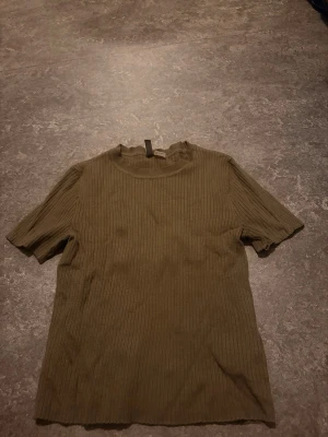 Olivgrön ribbad topp med kort ärm - Säljer en olivgrön ribbad topp med korta ärmar och hög hals. Toppen har en enkel, stilren design och är perfekt att matcha med både jeans och kjol. Materialet känns mjukt och stretchigt för en bekväm passform.