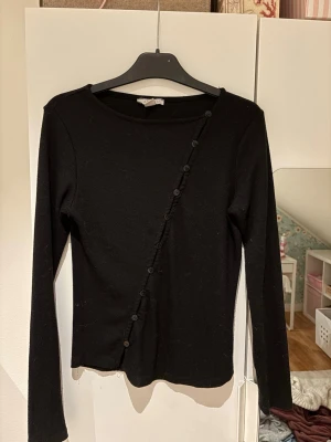 Svart  topp med knappar - Snygg svart super trendig långärmad topp med ribbad struktur och dekorativa knappar i diagonal linje framtill. Toppen har en tight passform och rund halsringning, perfekt att styla med jeans eller kjol.