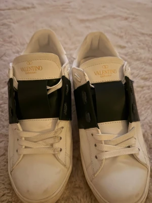 Valentino Garavani vita sneakers - Snygga vita sneakers från Valentino Garavani med bred svart läderdetalj över snörningen och guldfärgad logga på plösen. Skorna har klassisk låg profil och vita skosnören. Perfekta för dig som vill ha en stilren och exklusiv look.