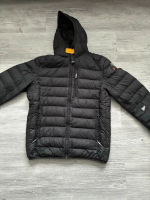 Svart dunjacka med huva Parajumpers - Svart dunjacka från Parajumpers med huva och dragkedja framtill. Jackan har två sidofickor med dragkedja och en bröstficka. Klassisk quiltad design och patch på ärmen. Perfekt för kalla vinterdagar. Lite söndrig vid armen men det är inget som inte går att fixa.
