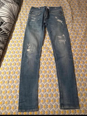 Replay Anbass blå slitna jeans - Säljer ett par Replay Anbass jeans i ljusblå tvätt med slitningar och hål på benen för en riktigt snygg, avslappnad look. Jeansen har klassisk femficksdesign, normal passform och är gjorda i hållbar bomull. Perfekta för dig som gillar streetstyle och vill ha något bekvämt.