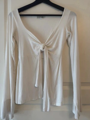 Vit knytblus från Design by Si - Supersnygg vit blus från Design by Si med lång ärm och djup v-ringning. Toppen har en knytning framtill som ger en trendig och feminin look. Perfekt att styla med jeans eller kjol för en chic outfit. Bra skick.  Storlek xs/s 