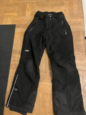 Svarta täckbyxor från North Bend - Svarta täckbyxor från North Bend med dragkedjeförsedda fickor och förstärkta knän. Byxorna har bred passform, reflexdetaljer och är perfekta för vinterns alla äventyr. Materialet är slitstarkt och vattenavvisande.
