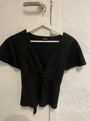 Svart omlottopp med knyt  - Snygg svart omlottopp från Gina  med djup v-ringning och korta, vida ärmar. Toppen har knytband i midjan för en justerbar passform och är tillverkad i mjukt bomullsmaterial. Perfekt att styla med jeans eller kjol.💞