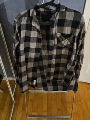 Rutig svart och grå skjorta  - Snygg långärmad skjorta i svart och grått rutigt mönster. Skjortan har klassisk krage, knäppning framtill och en bröstficka. Perfekt för en avslappnad och trendig look.