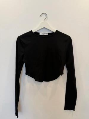 Svart croppad topp från NA-KD - Svart långärmad croppad topp från NA-KD med rund halsringning. Tillverkad i ribbad bomull för en bekväm och trendig look. Perfekt att matcha med höga jeans eller kjol.