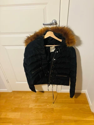 Moncler jacka - Svart dunjacka med huva och brun fuskpäls runt kanten. Storlek XS till S. Jackan har dragkedja, snörning i midjan och två snedställda fickor med dragkedja framtill. Perfekt för kalla vinterdagar och har en modern, kortare passform. Äkta solklart, så intresserad hojta till bara 😃