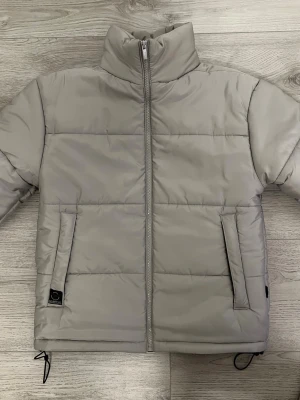 Ljusgrå pufferjacka med hög krage - Snygg silvergrå kort pufferjacka med ståkrage och raka linjer från Le Breve. Jackan har två fickor framtill med diskret logga, dragkedja hela vägen och justerbara snören vid nederkanten. Perfekt för kalla dagar och enkel att matcha med din stil.