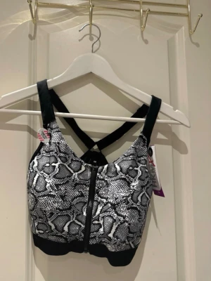 Sport-bh med ormmönster och dragkedja - Hunkemöller sport-bh med svartvitt ormmönster framtill och svarta detaljer. Bh:n har breda axelband som korsas i ryggen och en dragkedja framtill. Materialet är stretchigt och bekvämt, perfekt för träning eller en aktiv vardag.