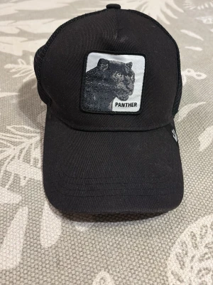 Svart truckerkeps med pantermärke - Svart truckerkeps med mesh på sidorna och bak, samt ett broderat märke framtill med en svart panter och texten 'PANTHER'. Justerbar passform baktill. Perfekt för dig som gillar djurmotiv och streetstyle.