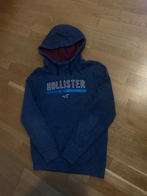 Blå Hollister hoodie med tryck - Mörkblå hoodie från Hollister med stor huva och dragsko. Framsidan har ett vitt och blått Hollister-tryck samt den klassiska loggan. Insidan av huvan är vinröd. Mjuk bomullsblandning och känguruficka framtill. Perfekt för en avslappnad stil.