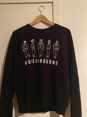 Svart Moschino Uomo sweatshirt - Cool svart sweatshirt från Moschino Uomo med vitt tryck av fem stiliserade figurer och texten MOSCHINOUOMO på bröstet. Tröjan har rund halsringning och är tillverkad i mjukt material, perfekt för en trendig och avslappnad look.