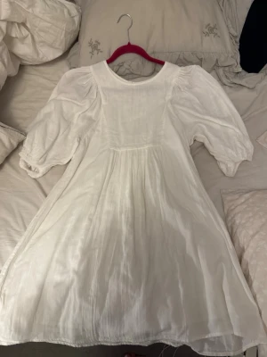 Vit kort klänning med puffärm  - Supersöt vit klänning med puffärmar och snörning i ryggen. Klänningen har en luftig passform, rund halsringning och är tillverkad i ett lätt tyg som känns perfekt för varmare dagar. Passar dig som gillar romantisk och trendig stil. Perfekt till studenten 