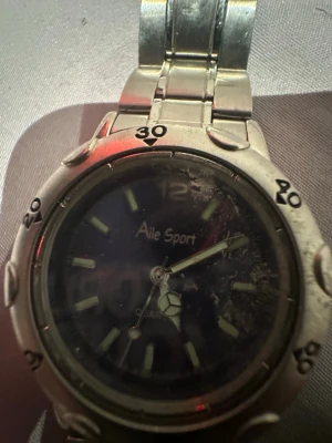 Aile Sport silverfärgad klocka - Stilren klocka från Aile Sport med silverfärgat metallarmband och mörk urtavla. Klockan har tydliga siffror och markeringar runt boetten samt klassisk länkdesign. Perfekt accessoar för dig som gillar sportig och tidlös stil. Funkar ej!