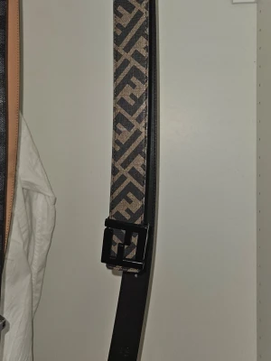 Fendiii svart och beige skärp - Säljer ett Fendiii skärp i svart skinn med beige och svart Fendi-logomönster på framsidan. Skärpet har en stilren svart metallspänne med Fendi-logga och präglad text på insidan. Perfekt accessoar för att lyfta din outfit.