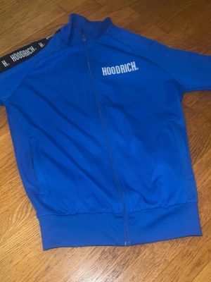 Blå zip hoodie från Hoodrich - Snygg blå hoodie från Hoodrich med dragkedja framtill och vita logotexter på bröstet och längs ärmarna. Hoodien har ribbade muddar och två fickor på sidorna. Perfekt för en streetwear-look och riktigt bekväm att bära.