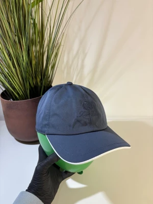 Loro Piana Keps - Snygg mörkblå keps från Lord Lane med broderad logga framtill. Kepsen har böjd skärm med vit kant och klassisk sexpanelsdesign. Perfekt accessoar för en sportig och stilren look.