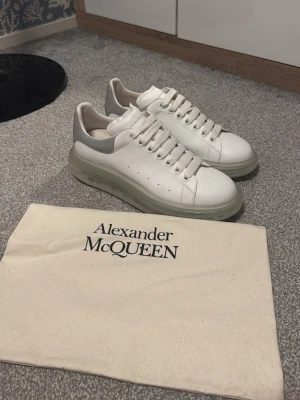 Alexander McQueen vita sneakers - Stilrena vita sneakers från Alexander McQueen med grå detalj på hälen och tjock, transparent sula. Skorna har klassisk snörning och är tillverkade i läder med perforerade sidor för extra stil. Perfekta för dig som vill ha en clean och modern look.