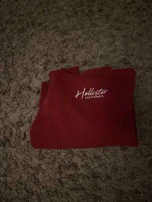 Vinröd hoodie från Hollister - Snygg vinröd hoodie från Hollister med vit broderad logga på bröstet och huvan. Klassisk känguruficka framtill och mjukt material som känns skönt mot huden. Perfekt för en avslappnad och trendig stil.